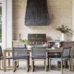 Modern custom outdoor kitchen and dining area in Fort Worth with built-in grill, stone wall, and sleek patio furniture.